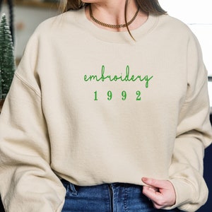 Puede incluir: Una sudadera beige con bordado verde que dice "embroidery 1992".
