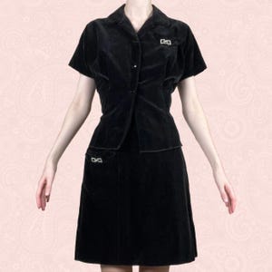 May include: A vintage black velvet two-piece set. The set includes a short-sleeved button-up shirt and a knee-length skirt. Both pieces feature decorative silver-toned accents. The outfit is displayed against a pink patterned background.