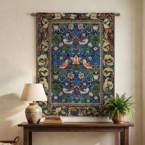 Puede incluir: Un tapiz detallado con un fondo azul y un diseño floral y de pájaros cuelga sobre una consola de madera oscura. Una lámpara, libros y un helecho en maceta están sobre la mesa. Una cesta y una alfombra tejidas están en primer plano.