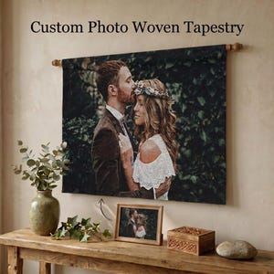 May include: A custom photo woven tapestry hanging on a wall, featuring a couple in an embrace against a dark green foliage background. The text Custom Photo Woven Tapestry is printed above the tapestry, which is suspended from a wooden rod.