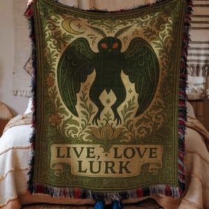 May include: A green and gold throw blanket featuring a dark silhouette of a moth-like creature with large wings. The blanket has a decorative border and the text "LIVE, LOVE, LURK". The blanket has a fringed edge.
