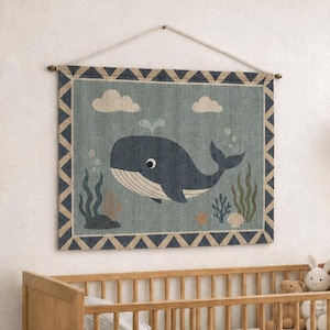 May include: A rectangular wall hanging featuring a blue whale illustration on a textured background with a geometric border, suspended by a string above a wooden crib with white bedding, a plush toy, and a mobile hanging nearby.