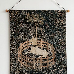 Könnte beinhalten: Ein rechteckiger Wandbehang, der ein weißes Einhorn zeigt, das in einem Holzzaun ruht. Der Hintergrund ist ein dunkler, dichter Wald voller verschiedener Blumen und Pflanzen. Der Wandbehang ist an einer Holzstange mit einer dunklen Schnur befestigt.