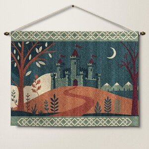 May include: A rectangular wall hanging featuring a castle on a hill under a crescent moon. The scene includes a winding path, trees with orange and red leaves, and distant mountains. The design is framed by a decorative border with a white knot pattern.
