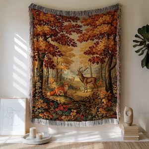 May include: Woven tapestry throw blanket featuring a forest scene with a deer and fox. The blanket has a fringed edge and depicts a colorful autumn landscape with trees, foliage, and mushrooms. The color palette includes shades of brown, orange, and green.