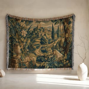 Op de afbeelding: Geweven wandtapijt deken met een landschap met een kronkelende weg, kasteel en bomen. De deken heeft een franjerand en is in tinten groen, blauw en beige. Een grote urn met bloemen staat op de voorgrond.