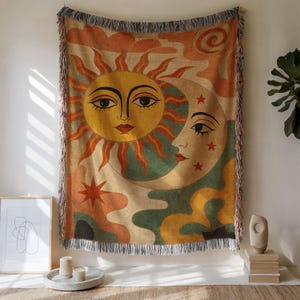 May include: Woven tapestry featuring a sun and moon design with faces, stars, and abstract shapes in orange, yellow, green, and beige. The tapestry has a fringed edge and is displayed on a wall.