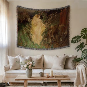 May include: A woven tapestry featuring a classical painting of two figures in flowing gowns amidst a lush, green landscape with white flowers. The tapestry is rectangular with a fringed border and is displayed above a white sofa with neutral-toned pillows.
