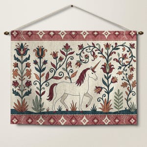 Puede incluir: Un tapiz rectangular de tela que presenta un unicornio blanco central con crin y cuerno de color granate. El diseño incluye motivos florales estilizados en tonos granate, verde azulado y verde salvia sobre un fondo crema, enmarcado por un borde geométrico granate.