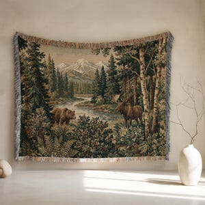 May include: A woven tapestry throw blanket featuring a detailed landscape scene with a brown bear and a moose near a river. The background displays snow-capped mountains, dense pine forests, and birch trees. The edges have a decorative fringe.