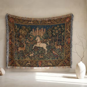 Puede incluir: Un tapiz tejido que representa una escena de bosque medieval con un unicornio blanco, ciervos, conejos y un castillo. El tapiz tiene un fondo azul con bordes ornamentados y flecos.