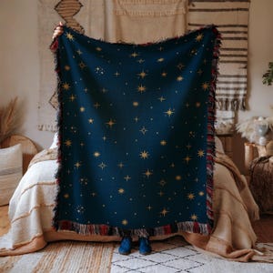 May include: A navy blue throw blanket with a gold star pattern. The blanket has a fringed edge with red and gold accents. The blanket is held up in front of a bed with a neutral-toned bedspread.