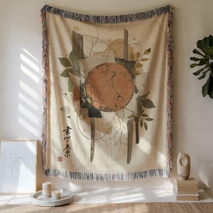 May include: Beige woven throw blanket with an abstract design. It features a cracked orange circle, wooden beams, and green leaves. The blanket has a fringed edge and Japanese text.