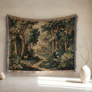 Puede incluir: Un tapiz de pared tejido rectangular que presenta un paisaje forestal con árboles altos, follaje verde denso y un camino de tierra que serpentea por el centro. Un pequeño ciervo se encuentra en el lado izquierdo del camino. Borde con flecos.