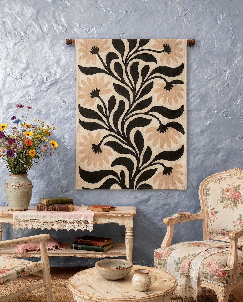 May include: A rectangular wall hanging featuring a repeating pattern of large, light beige flowers with dark centers and bold, black, flowing, leaf-like stems on a cream-colored fabric background, suspended from a dark wooden rod.