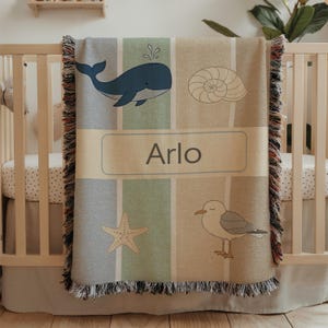 May include: A personalized baby blanket with a nautical theme. The blanket features a blue whale, seashell, starfish, and seagull design. The name "Arlo" is displayed in a rectangular box. The blanket is draped over a crib.