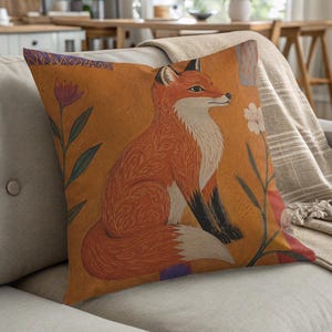 May include: A square throw pillow featuring a vibrant orange background with a central illustration of a sitting fox. The fox has a white chest and tail tip with black paws. The design includes stylized floral elements in red and white.