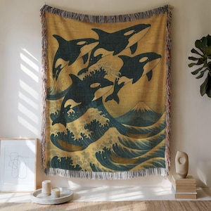May include: A woven throw blanket featuring a stylized illustration of orcas leaping from ocean waves, with a mountain in the background. The design is in shades of blue, gold, and cream, with a fringed edge.