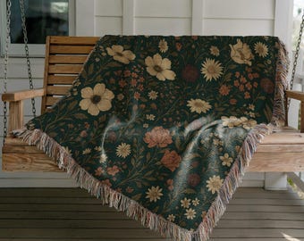 May include: A dark green floral throw blanket with a botanical pattern of beige, peach, and brown flowers. The blanket has a fringed edge and is draped over a wooden porch swing. The background includes a white house and a window.