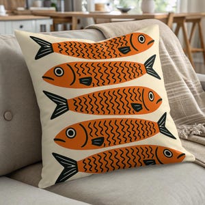 Peut inclure: Un coussin décoratif avec un motif répété de poissons orange avec des détails noirs sur un fond crème. Les poissons ont un design stylisé avec des motifs en zigzag et sont disposés verticalement. Le coussin est posé sur un canapé clair.