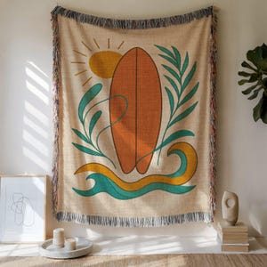 Op de afbeelding: Een beige geweven sprei met een surfboard ontwerp. Het ontwerp toont een oranje surfboard, een gele zon, turquoise golven en groen gebladerte. De sprei heeft een gefranjerde rand en wordt binnenshuis getoond.