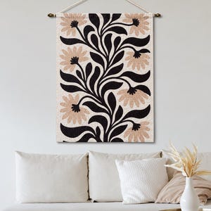 May include: A rectangular wall hanging featuring a repeating botanical pattern of black leaves and light tan flowers on a cream-colored fabric background. The textile is suspended from a horizontal rod with a white cord for mounting on a wall.