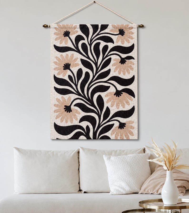 May include: A rectangular wall hanging featuring a repeating botanical pattern of black leaves and light tan flowers on a cream-colored fabric background. The textile is suspended from a horizontal rod with a white cord for mounting on a wall.