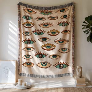 May include: Beige throw blanket with a repeating pattern of stylized eyes in teal, orange, and brown. The blanket has a fringed edge in shades of gray, red, and beige. The blanket is hanging on a wall.