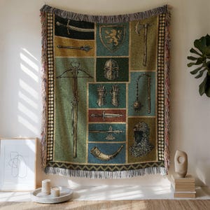 May include: Woven tapestry blanket featuring a medieval theme. The design includes illustrations of swords, shields, helmets, and other weaponry. The color palette consists of greens, blues, and browns, with a fringed edge. The blanket is displayed on a wall.