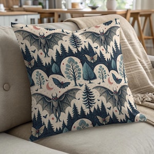May include: Square decorative pillow with a repeating pattern of bats, trees, and crescent moons in shades of blue, gray, and cream. The pillow is placed on a beige sofa with a matching throw blanket. The pillow's design has a whimsical, gothic theme.