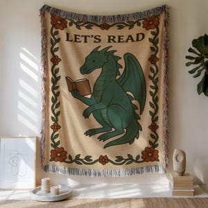 Puede incluir: Manta tejida beige con un dragón verde leyendo un libro, con el texto "LET'S READ". La manta tiene un borde de vid floral y bordes con flecos. Un artículo decorativo para un rincón de lectura.