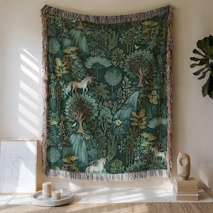 Pode incluir: Uma manta de tapeçaria com uma cena de floresta com cachoeiras, árvores e unicórnios em tons de verde, bege e branco. A manta tem uma borda com franjas e está pendurada em uma parede.