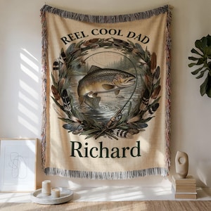 Puede incluir: Manta tejida beige con un diseño de pesca. La manta presenta una ilustración detallada de un pez, una caña de pescar y el texto "REEL COOL DAD" y "Richard". Los bordes están deshilachados.