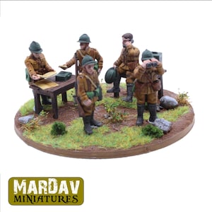 Könnte beinhalten: Fünf bemalte Miniaturfiguren von Soldaten in braunen und grünen Uniformen, die um einen Holztisch mit einer Karte darauf stehen. Die Figuren sind in einer militärischen Umgebung positioniert, wobei ein Soldat durch ein Fernglas schaut. Die Figuren stehen auf einem runden Sockel mit grünem Gras und braunem Schmutz.