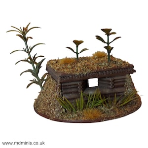 Può includere: Un bunker in miniatura di legno con un tetto di terra e una piccola apertura per un'arma. Il bunker è circondato da piccole piante verdi ed erba marrone.