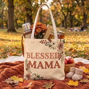 Könnte beinhalten: Eine cremefarbene Stofftasche mit dem Schriftzug BLESSED MAMA in großen braunen Buchstaben, verziert mit Blumenillustrationen in der oberen rechten und unteren linken Ecke. Die Tasche ist mit einer Thermoskanne, einem Buch und einem Glas gefüllt.