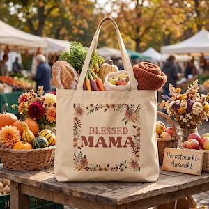 Könnte beinhalten: Eine beigefarbene Stofftasche mit dem Text BLESSED MAMA in braunen Buchstaben, umgeben von einem Blumenkranz. Die Tasche ist gefüllt mit einem Laib Brot, Karotten, einem Kürbis, Äpfeln und einem braunen Strickartikel auf einem Holztisch.