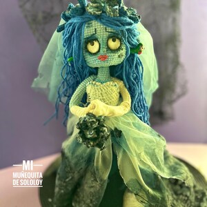 May include: A crocheted doll with light blue skin, large yellow eyes, and long blue yarn hair, wearing a white bodice and a flowing, layered green and blue fabric skirt. The doll holds a dark bouquet and wears a sheer veil, with text reading MI MUÑEQUITA DE SOLOLOY.
