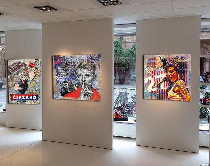Pu&ograve; includere: Tre opere d'arte incorniciate su una parete bianca. La parte pi&ugrave; a sinistra presenta un personaggio dei cartoni animati con una camicia a righe rosse e bianche e un cappello da marinaio blu. La parte centrale presenta un ritratto di David Bowie con una camicia rossa e un dito sulle labbra. La parte pi&ugrave; a destra presenta un ritratto di una donna con un microfono e uno sfondo rosso, bianco e blu.