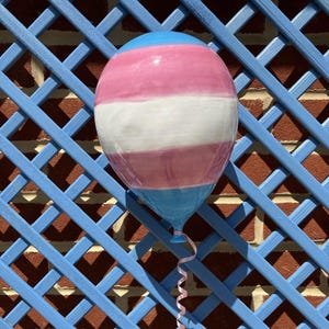 May include: A ceramic balloon in the colours of the transgender pride flag: light blue, pink, white, and pink. It is set against a blue lattice and a brick wall, with a pink ribbon attached.