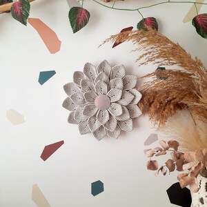 May include: A large, beige macrame flower wall hanging with a pink center. The flower is hanging on a white wall with a brown branch and green leaves in the background.