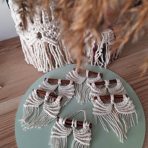 Pu&ograve; includere: Un set di sette arazzi da parete in macram&egrave; con bastoncini di legno. Ogni arazzo presenta un design in macram&eacute; bianco con frange. Gli arazzi sono esposti su una superficie verde chiaro.