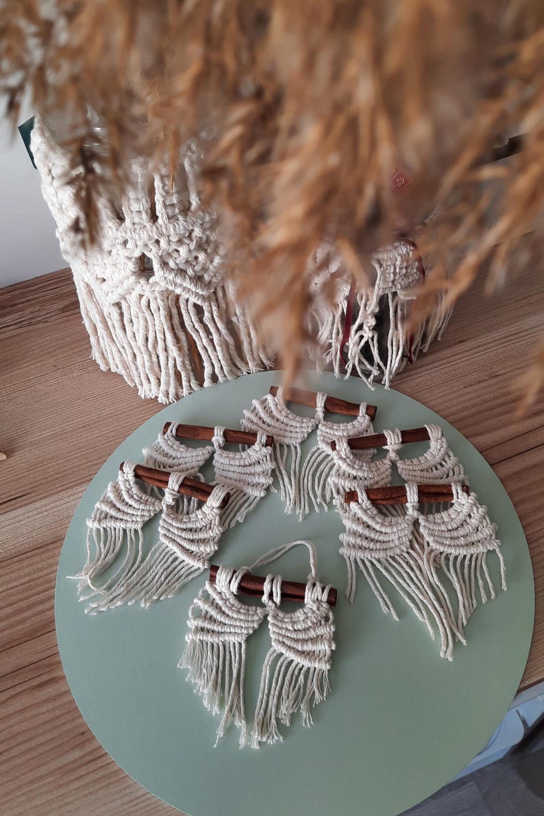 Pu&ograve; includere: Un set di sette arazzi da parete in macram&egrave; con bastoncini di legno. Ogni arazzo presenta un design in macram&eacute; bianco con frange. Gli arazzi sono esposti su una superficie verde chiaro.