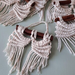 Pu&ograve; includere: Un set di cinque arazzi da parete in macram&egrave; con bastoncini di cannella. Ogni arazzo presenta un design geometrico con dettagli a frangia.