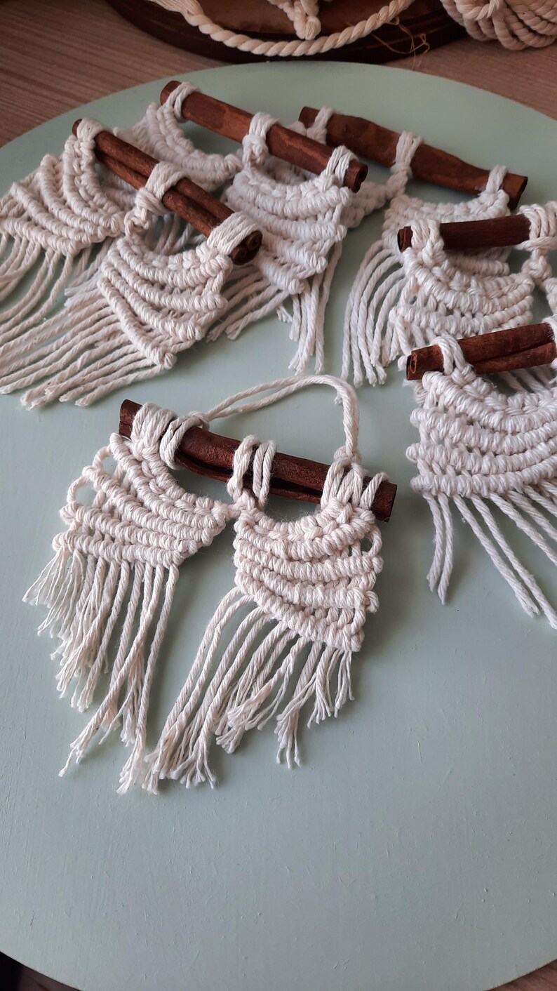 Pu&ograve; includere: Un set di cinque arazzi da parete in macram&egrave; con bastoncini di cannella. Ogni arazzo presenta un design geometrico con dettagli a frangia.