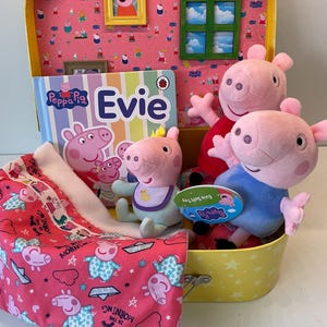 May include: A yellow suitcase containing Peppa Pig merchandise. The contents include plush pig toys, a book titled "Evie", and a pink patterned bag. The suitcase is decorated with Peppa Pig imagery and a small window.