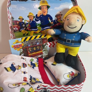 May include: A Fireman Sam themed gift set in a red and white chevron patterned tin. The set includes a Fireman Sam plush doll, a small pillow, and a shirt, all featuring Fireman Sam characters. The tin has a Fireman Sam scene on the inside.