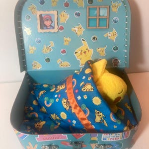 May include: A blue suitcase with a Pikachu theme. The suitcase is open and has a yellow Pikachu plush toy inside. The plush toy is sleeping under a blue blanket with a Pikachu pattern. The suitcase has a window and a picture of a character from the Pokemon cartoon.