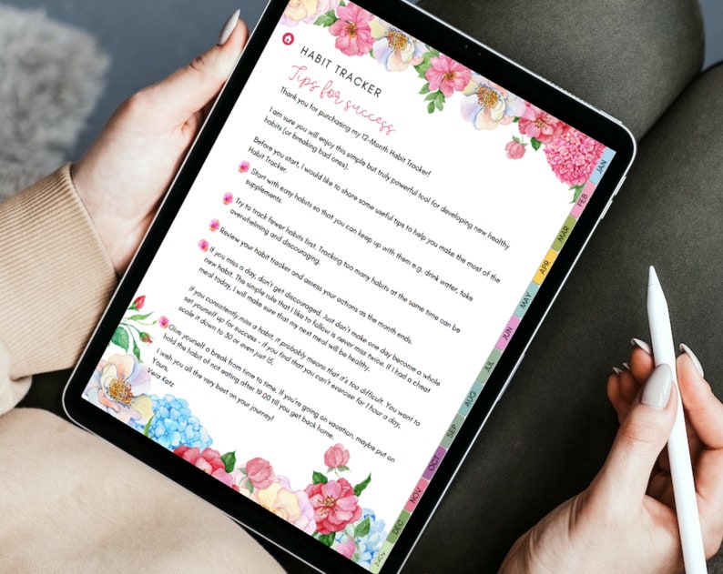 May include: A digital habit tracker displayed on a tablet, featuring floral watercolor accents. The screen shows text titled "Habit Tracker" with tips for success. The tablet is held by a person, with a stylus visible.
