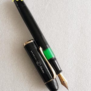 Puede incluir: Pluma estilográfica negra con plumín dorado y banda verde. El capuchón de la pluma está quitado, revelando el plumín. La pluma tiene el texto "Reopen Scholar" y "500" en amarillo.